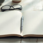 Open notebook, pen and reading glasses on a desk in soft daylight, suggesting careful study